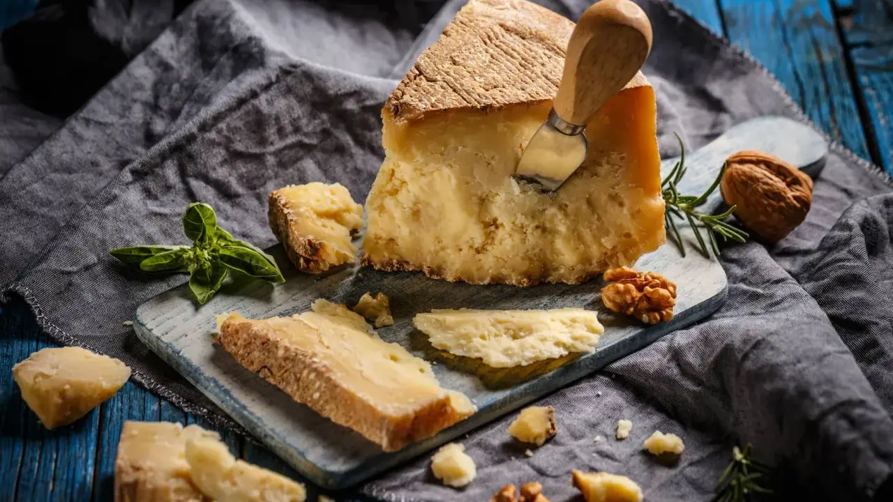 Kawałek parmezanu z nożem, okruchy sera, orzechy i bazylia. Czy parmezan można mrozić? Tak, można go mrozić, choć może stracić nieco na konsystencji.