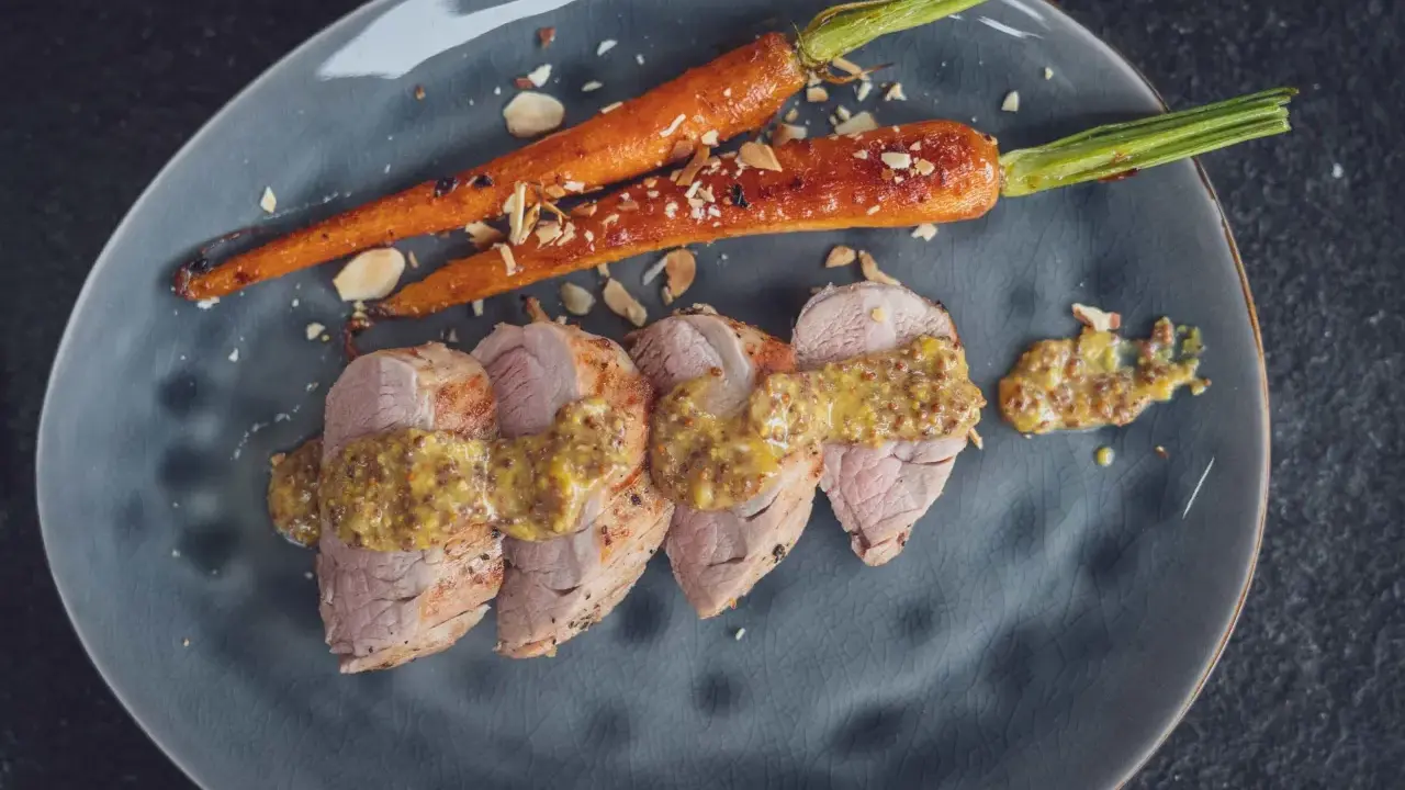 Zarte Scheiben von **schweinefilet grillen** mit Honig-Senf-Sauce und karamellisierten Karotten, bestreut mit Mandelsplittern.
