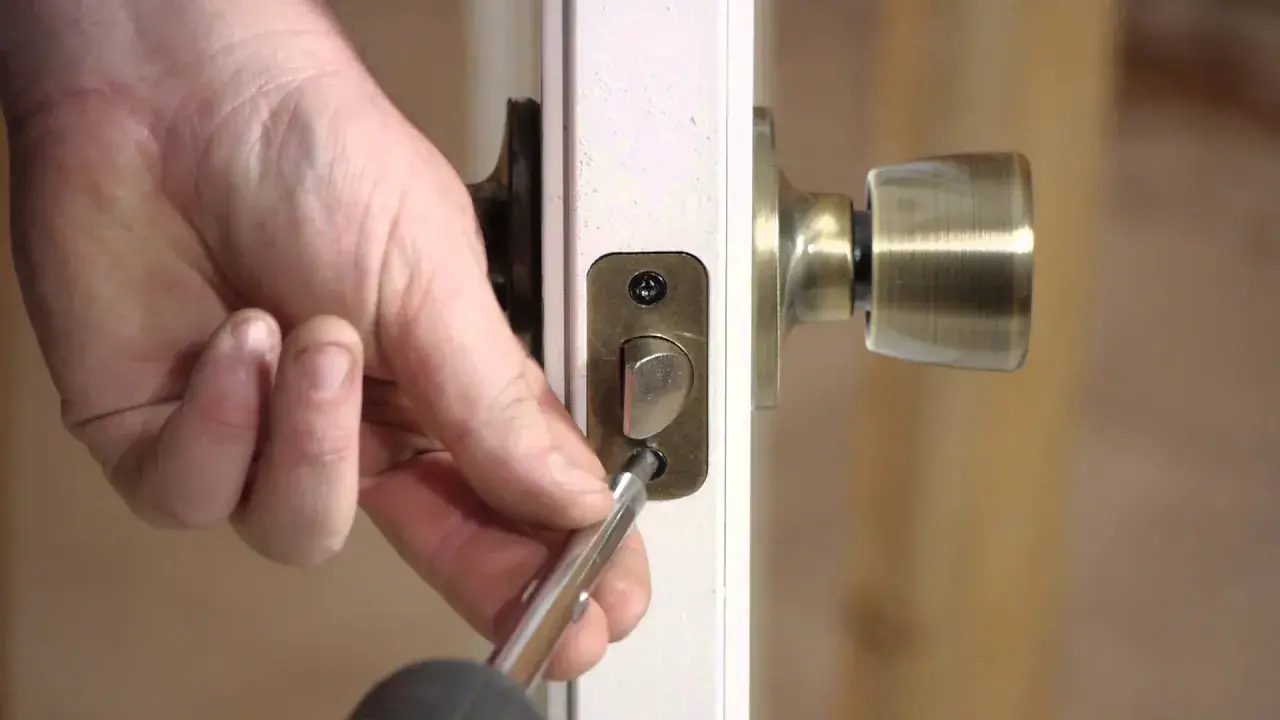 Mano usando un destornillador para desmontar una cerradura de puerta interior, quitando los tornillos de la placa.