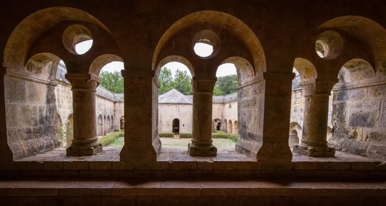 Vue à travers les arches de l'abbaye du Thoronet, un cloître en pierre avec un jardin verdoyant et des bâtiments anciens.