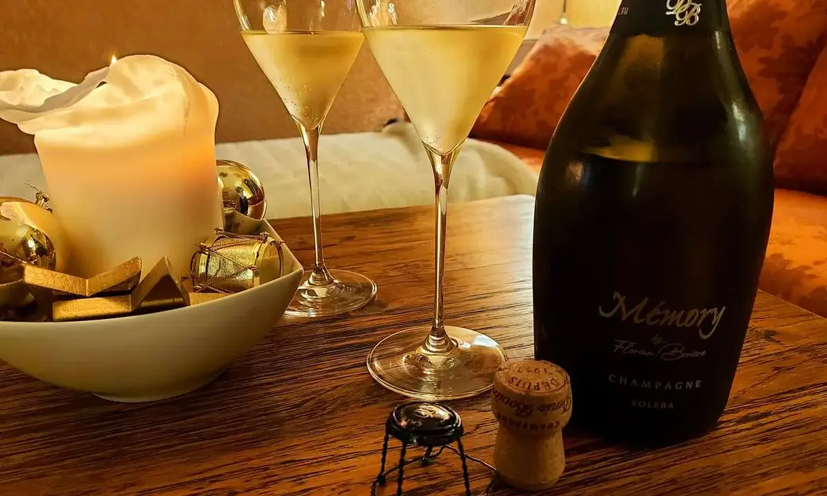 Zwei Gläser Champagner, eine Kerze und eine Flasche "Memory" auf einem Holztisch. Der Champagnergehalt ist hierbei nicht ersichtlich, aber die Stimmung ist festlich.