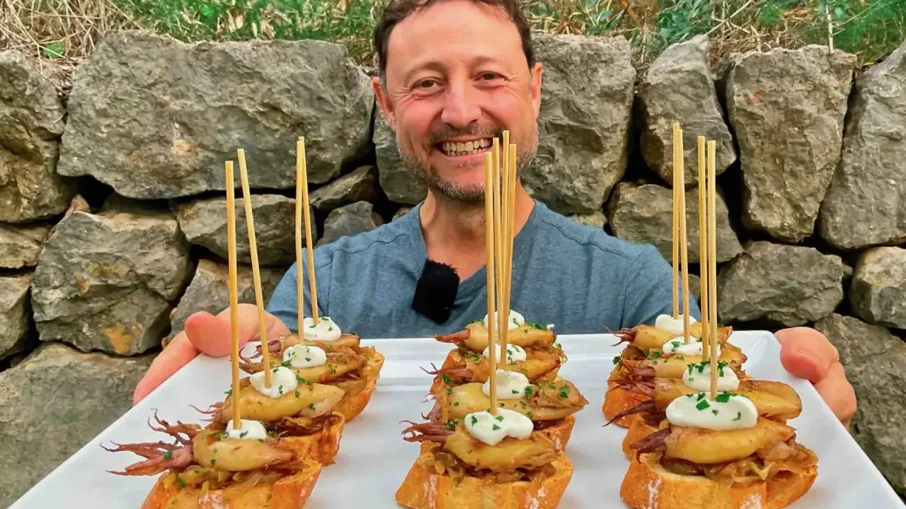 Hombre sonriente presenta cazuelitas de aperitivo con calamares y alioli sobre pan tostado, decoradas con brochetas.