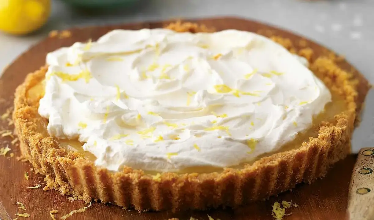 A lemon tart topped with whipped cream and lemon zest on a wooden board.