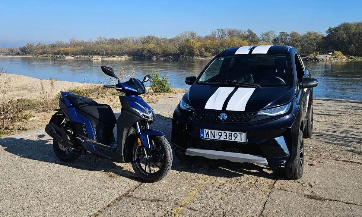 Niebieski skuter i czarne aixam auto z białymi pasami stoją nad rzeką.