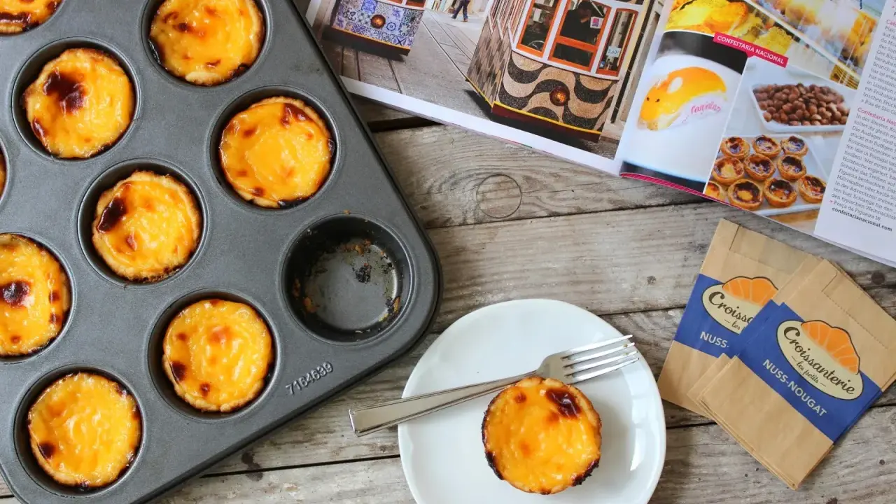 Pyszne, lekko przypieczone pastel de nata w blaszce i na talerzyku. Obok magazyn z widokiem na Lizbonę i tramwaje.