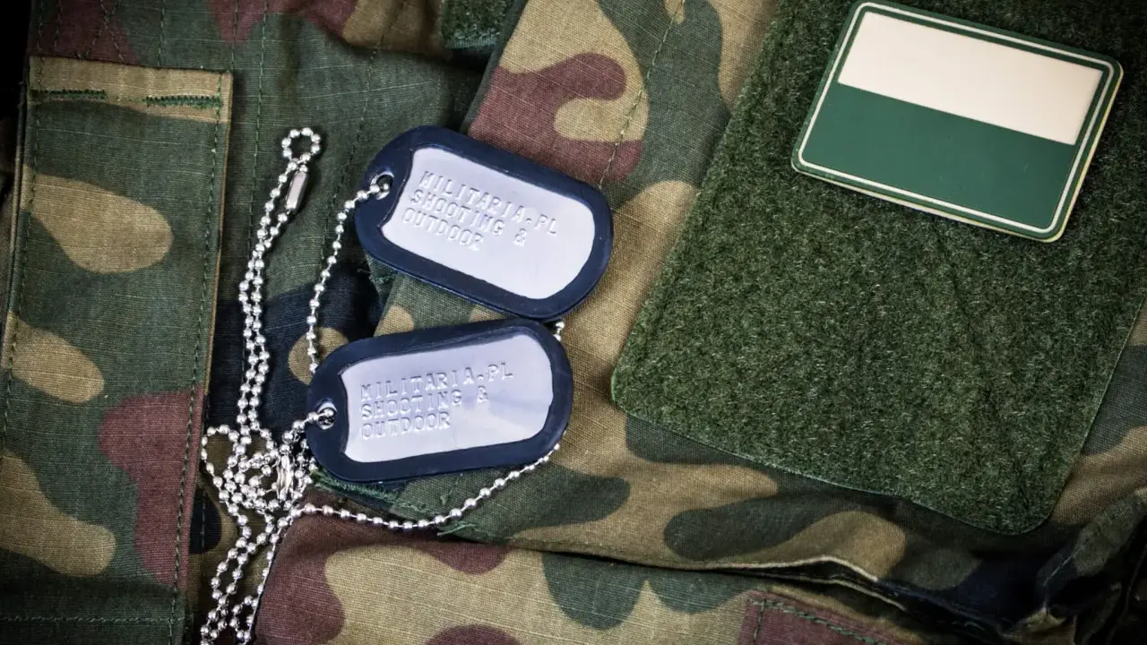Dwa nieśmiertelniki na mundurze moro. Jeden z nich zawiera napis "MILITARIA.PL SHOOTING & OUTDOOR".