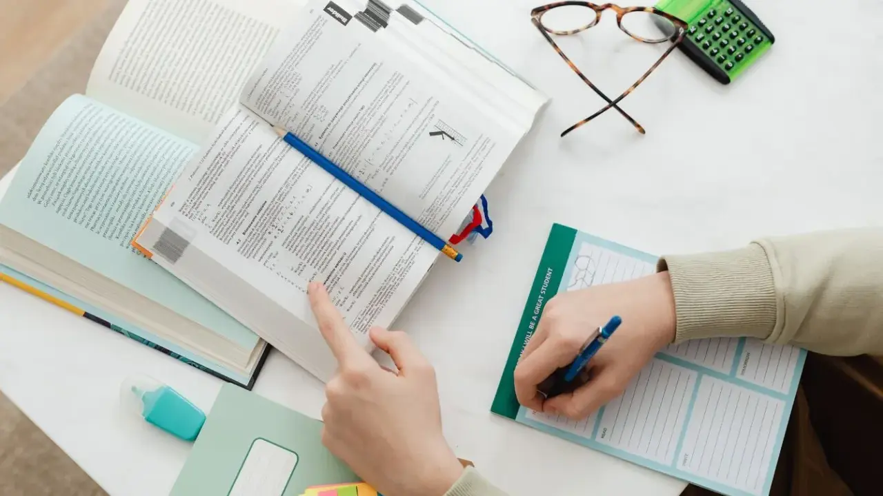 Uczeń pochyla się nad książkami, przygotowując się do egzaminu licencjackiego lub magisterskiego. Na stole leżą notatki, okulary i kalkulator.