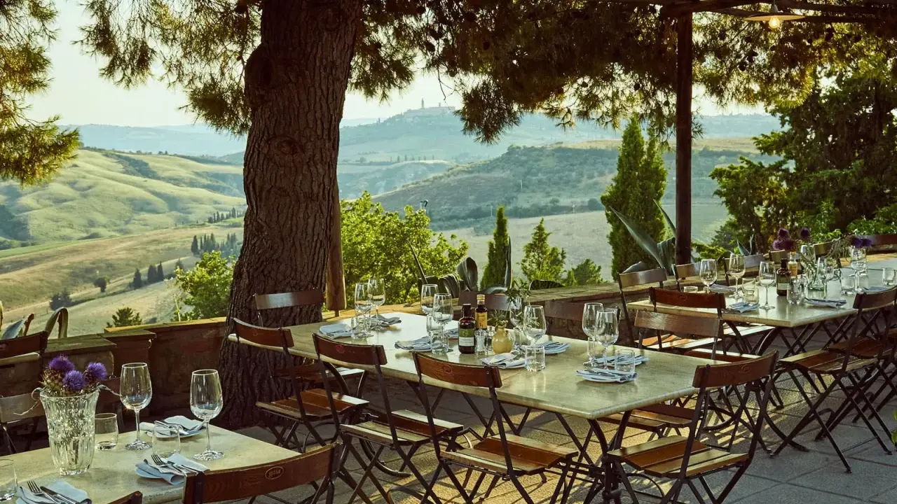 Tavoli apparecchiati sotto un pergolato con vista sulle dolci colline dell'entroterra toscana. Un invito a gustare la cucina locale.
