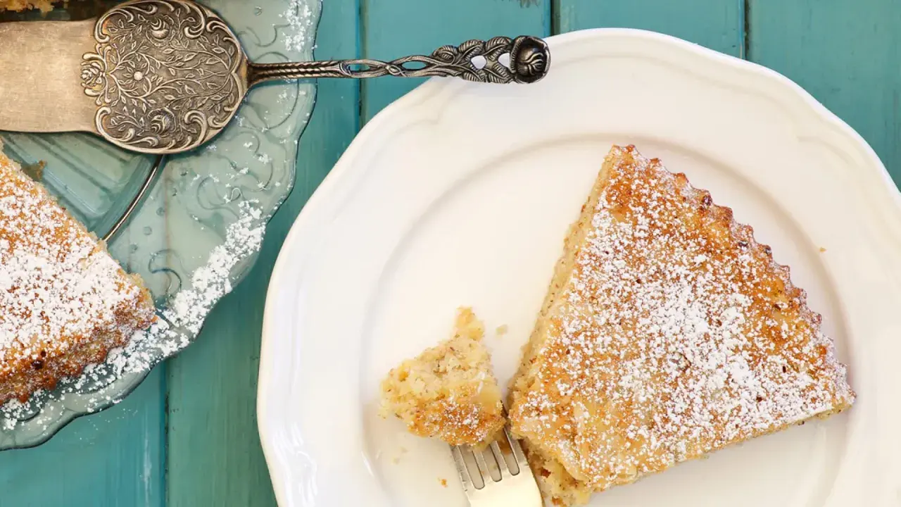 Saftige spanische Mandeltorte, bestäubt mit Puderzucker, auf einem weißen Teller. Ein Stück wurde bereits mit einer Gabel entnommen.