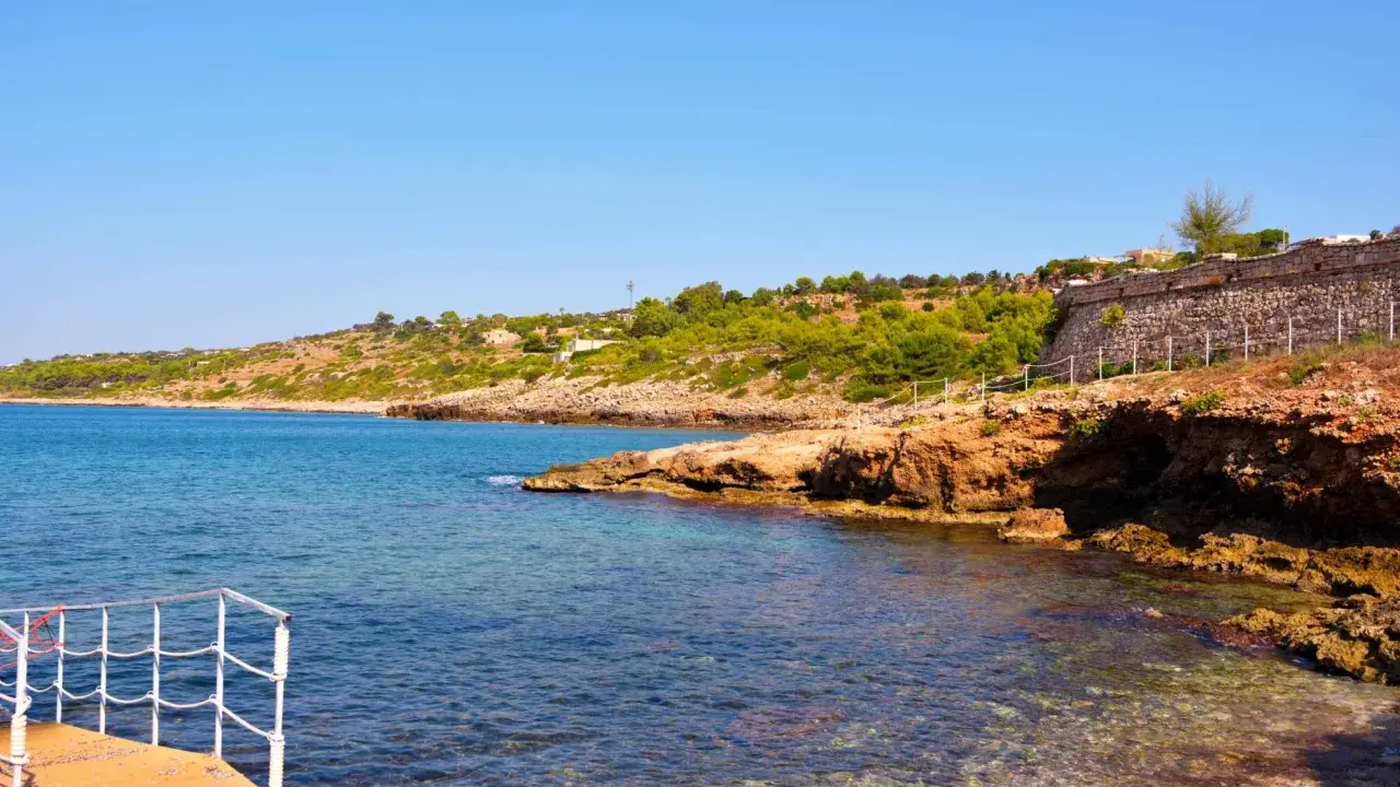 Scogliera rocciosa e mare cristallino a San Gregorio al mare, con vegetazione mediterranea e un muretto in pietra.