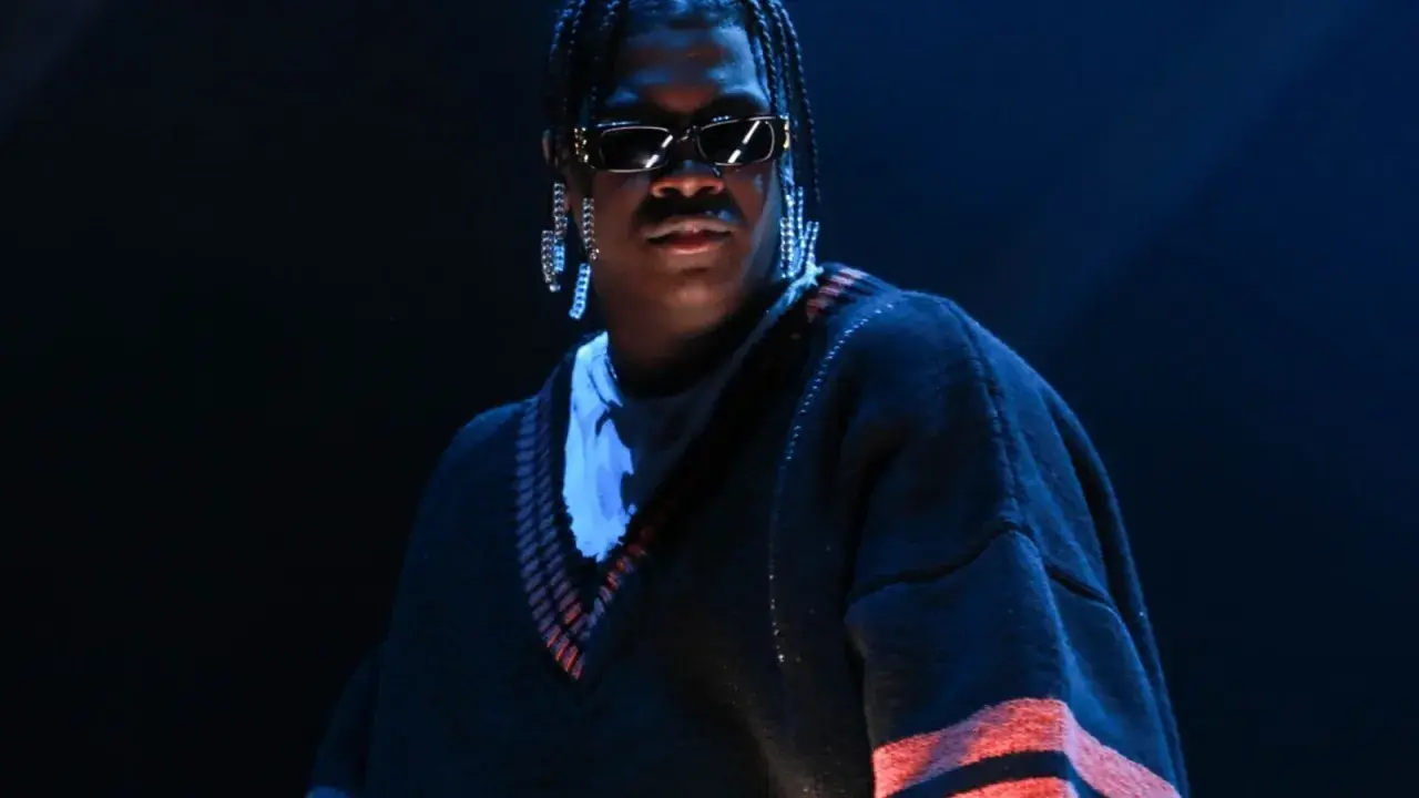 Lil Yachty w okularach i długich kolczykach, w swetrze z pomarańczowymi pasami. Let's start here.