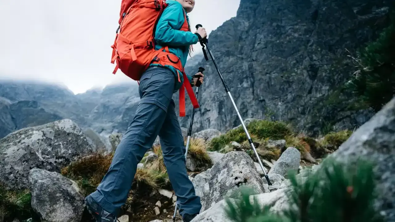 Kobieta z plecakiem wyrusza na wypad w góry, wspierając się kijkami trekkingowymi po kamienistym szlaku.