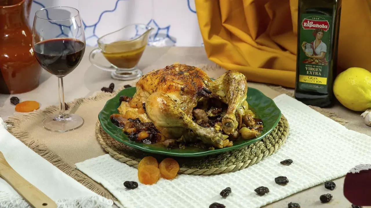 Pollo asado relleno, dorado y jugoso, servido en plato verde con pasas y orejones. Acompañado de vino tinto y aceite de oliva.