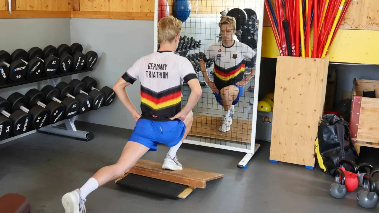 Ein Athlet im "Germany Triathlon"-Shirt macht Ausfallschritte im Athletik Training. Hanteln und Trainingsgeräte im Hintergrund.