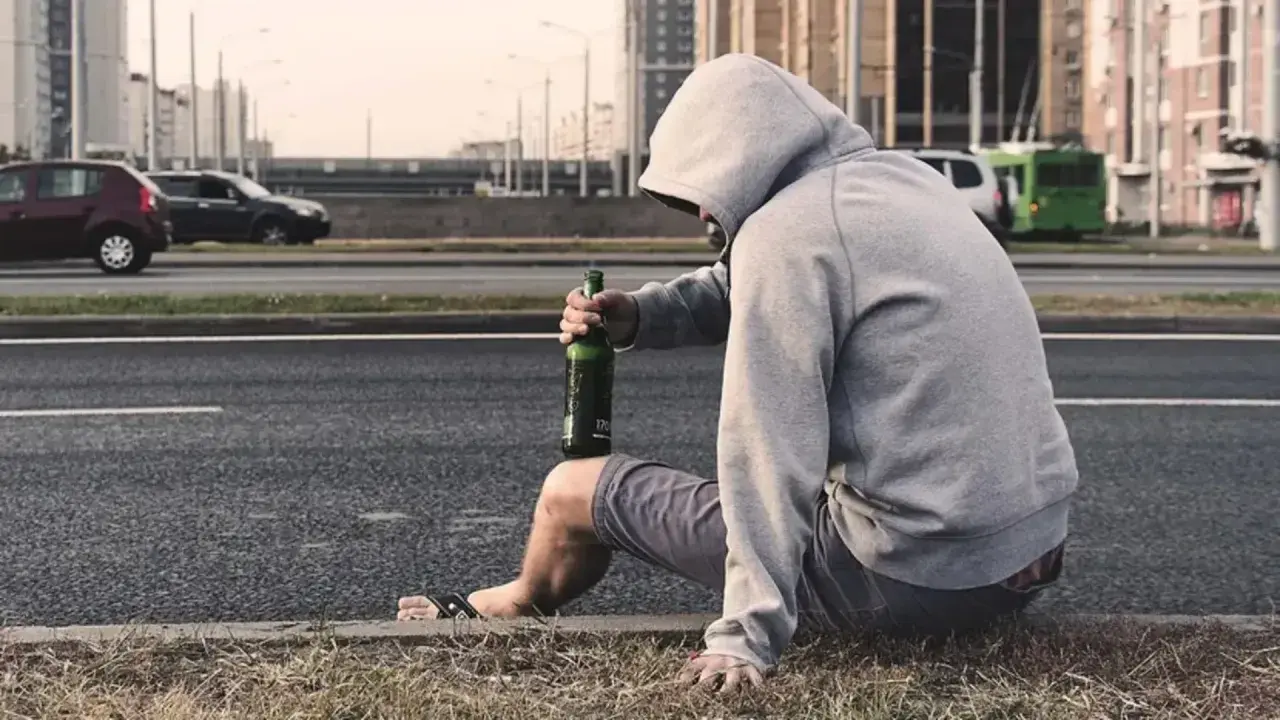 Mężczyzna w kapturze siedzi przy drodze z butelką piwa. Jego postawa sugeruje walkę z nałogiem, być może potrzebuje pomocy terapeuty od uzależnień.