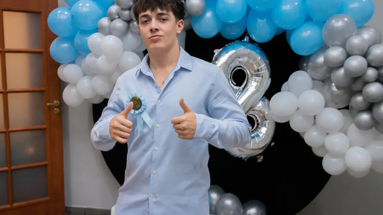 Chłopak w błękitnej koszuli z rozetką "18th Birthday" i balonami.
