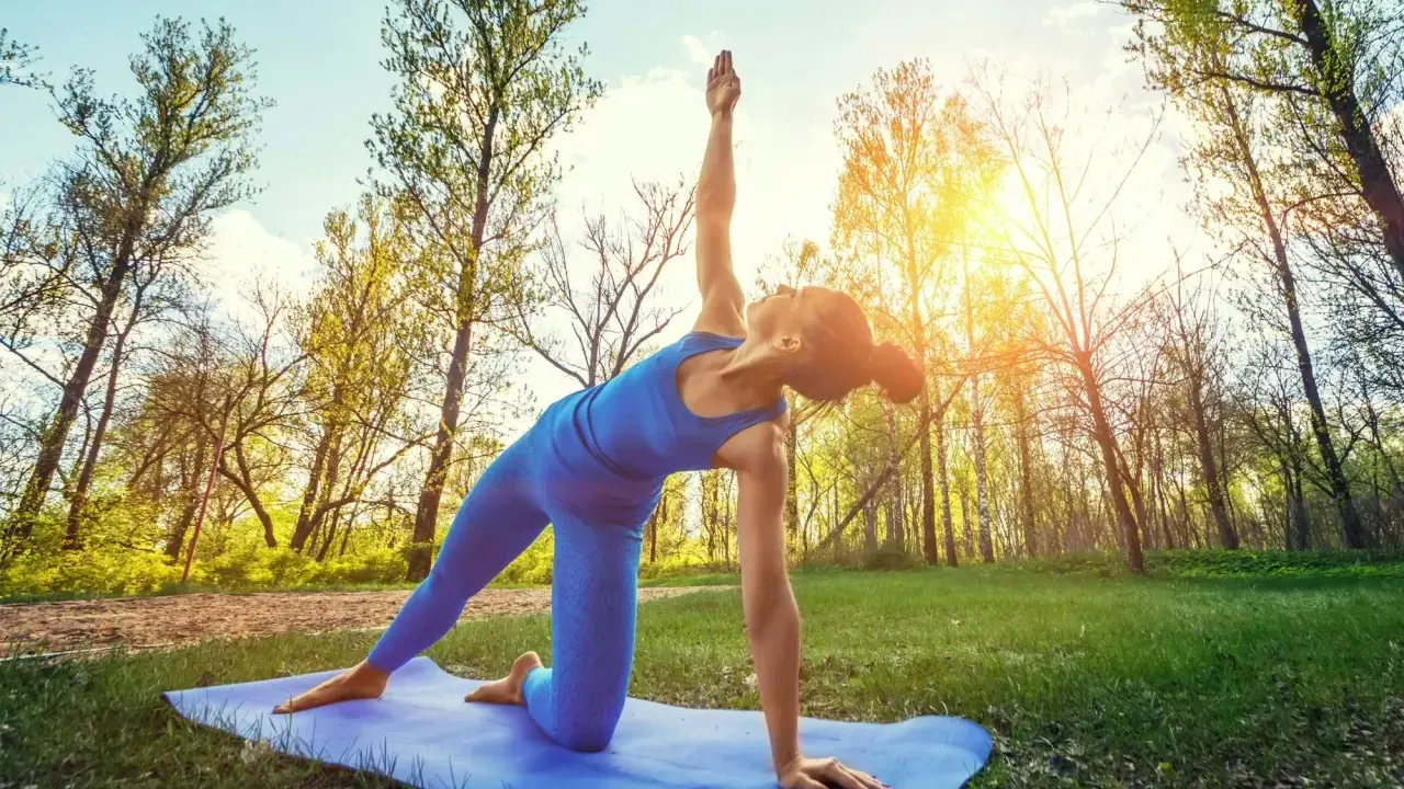 Kvinna utför yoga utomhus i en skogsmiljö. Solen skiner genom träden.