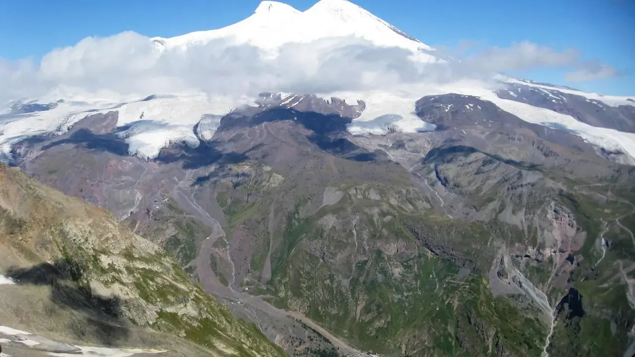 Majestatyczny Elbrus, najwyższy szczyt Europy, góruje nad skalistymi zboczami i lodowcami. W dole widać niewielką osadę, która pokazuje, gdzie leży Elbrus.