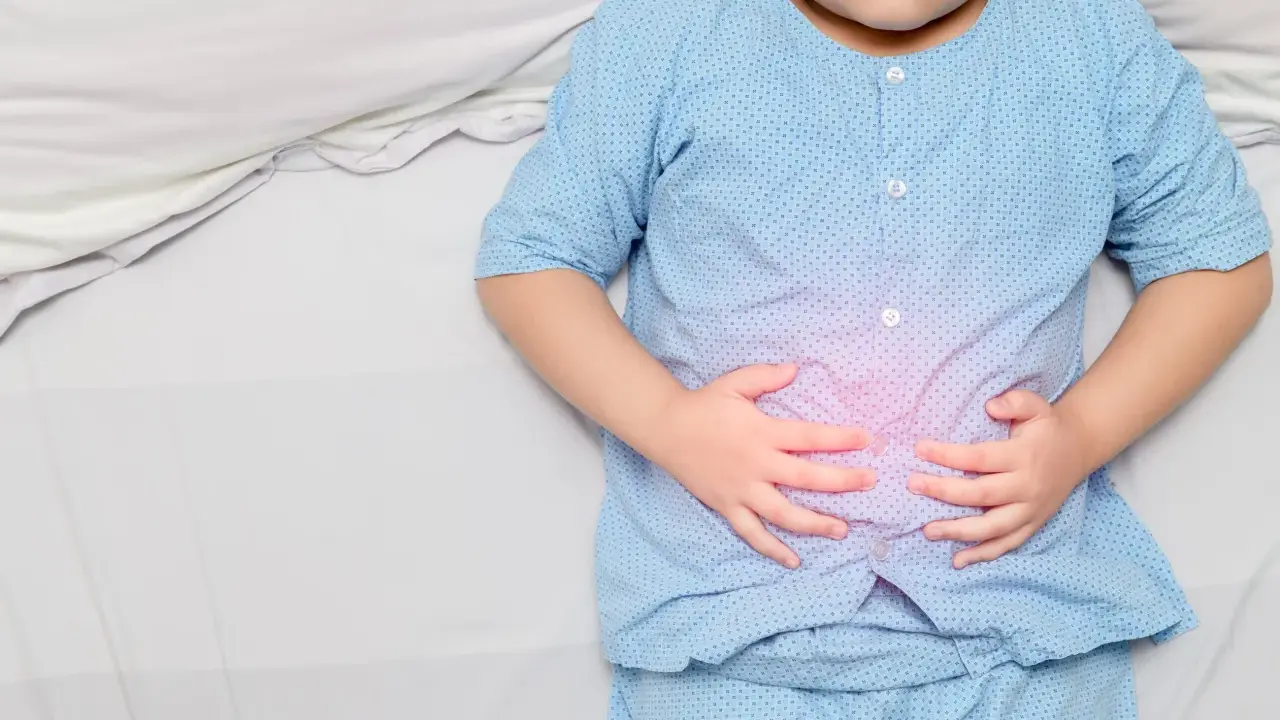Niño con pijama azul, acostado en cama blanca, se agarra la barriga con las manos. Se visualiza un área rosada en su abdomen, indicando dolor barriga niños.