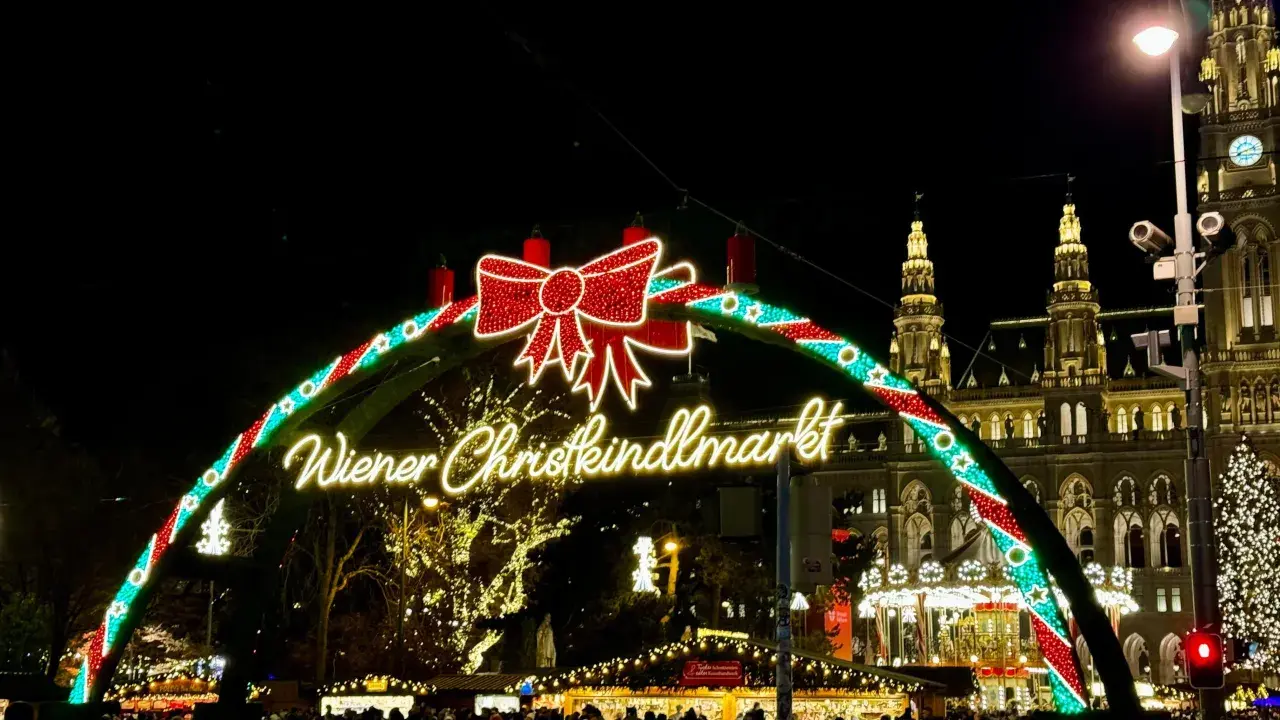 Wiedeński jarmark świąteczny w blasku świateł. Tłumy ludzi spacerują pod łukiem z napisem "Wiener Christkindlmarkt", w tle widać zabytkowe budynki i choinkę.