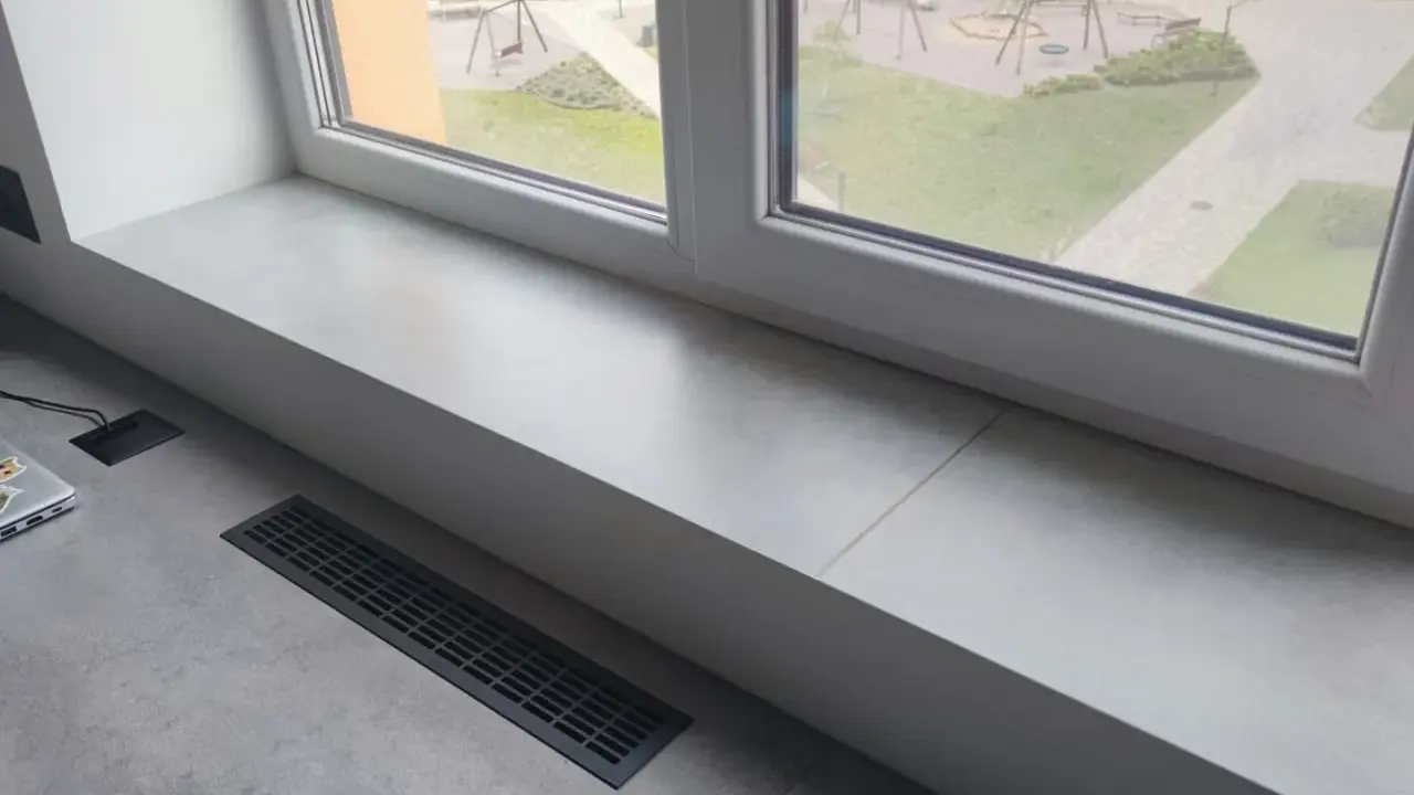 Jasny parapet z płytek wewnątrz, z widokiem na plac zabaw. Obok laptop i kratka wentylacyjna.