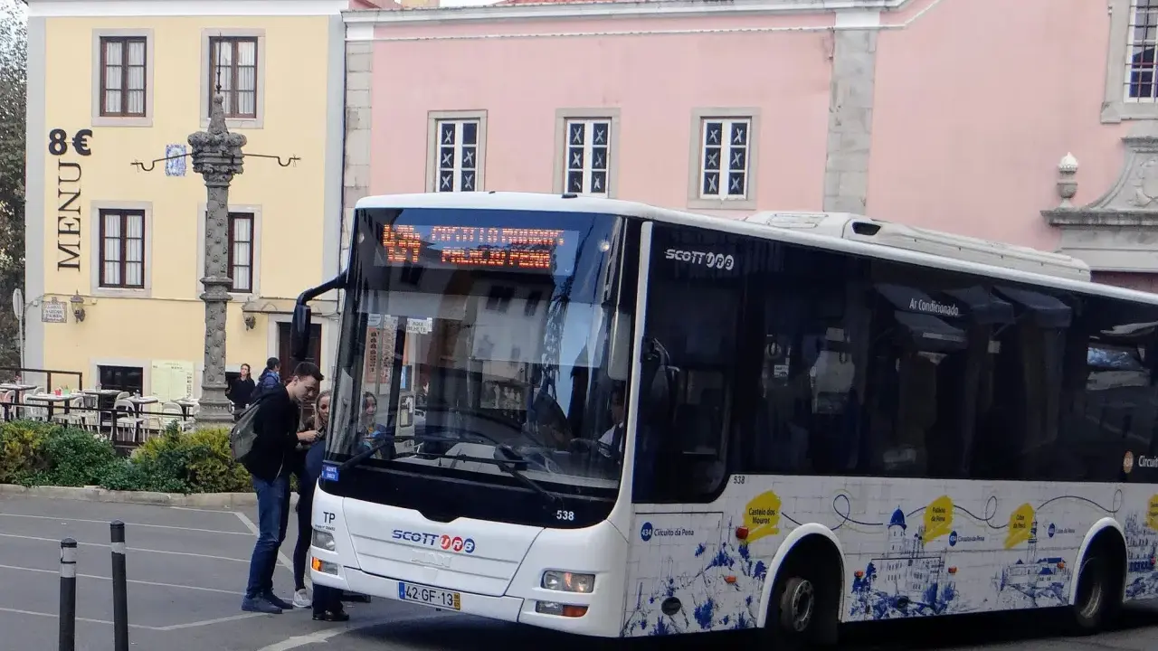 Ein Scott-Bus in Portugal hält an einer Haltestelle. Touristen steigen ein, während im Hintergrund bunte Gebäude und ein historischer Brunnen zu sehen sind.