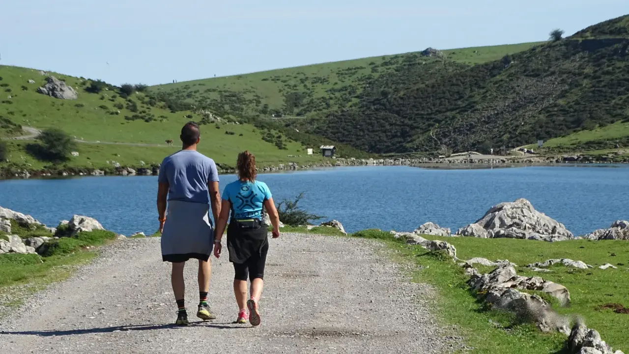 Ein Paar wandert auf einem Schotterweg am Ufer eines Sees. Grüne Hügel und Felsen prägen die **Spanien Natur**.