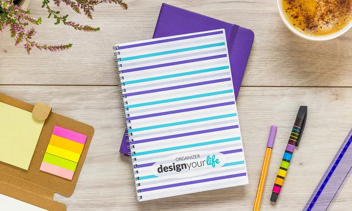 Pomysł na kalendarz: kolorowy notatnik z napisem "design your life", obok długopisy, karteczki i kawa.