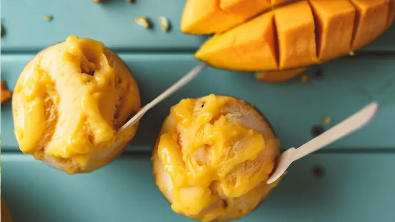 Jak zrobić sorbet? Dwa pucharki z mango sorbetem i kawałki świeżego mango na turkusowym tle.