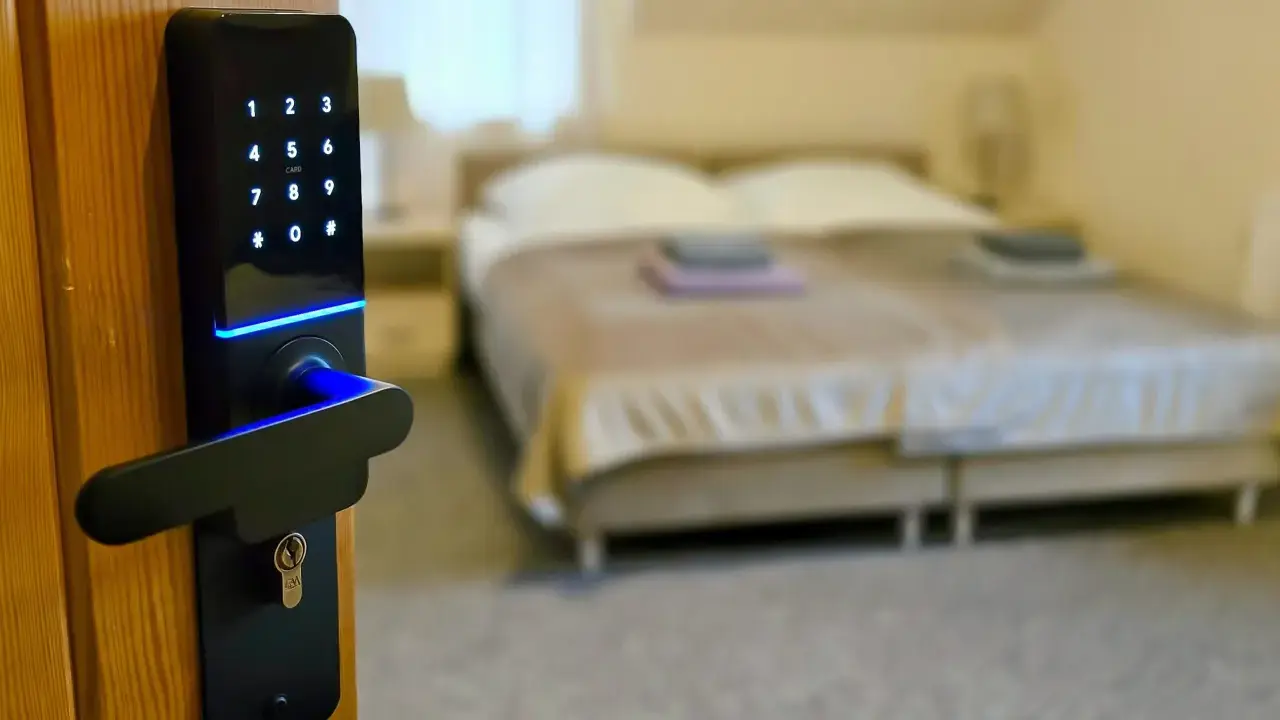 Cyfrowa klamka z klawiaturą numeryczną i niebieskim podświetleniem, symbolizująca digitalizację w hotelarstwie. W tle łóżko.