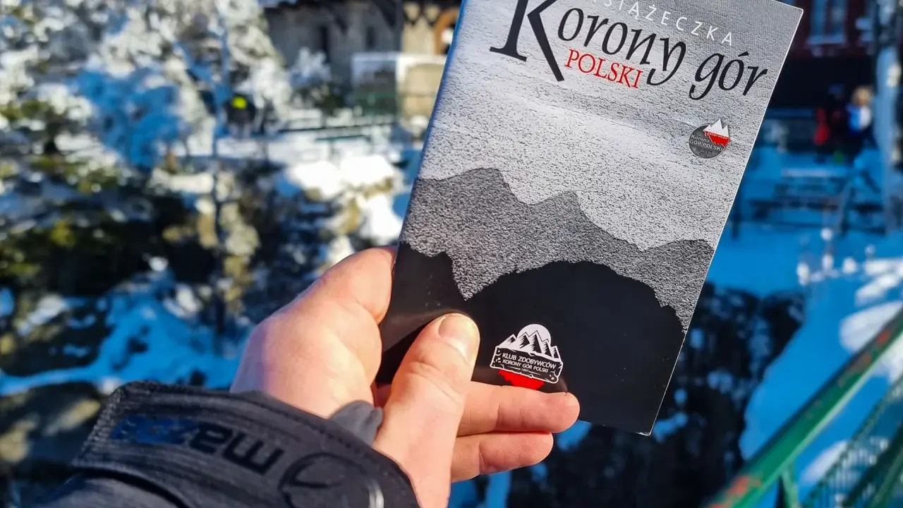 Dłoń trzyma książeczkę "Korona Gór Polski" na tle zimowego krajobrazu.
