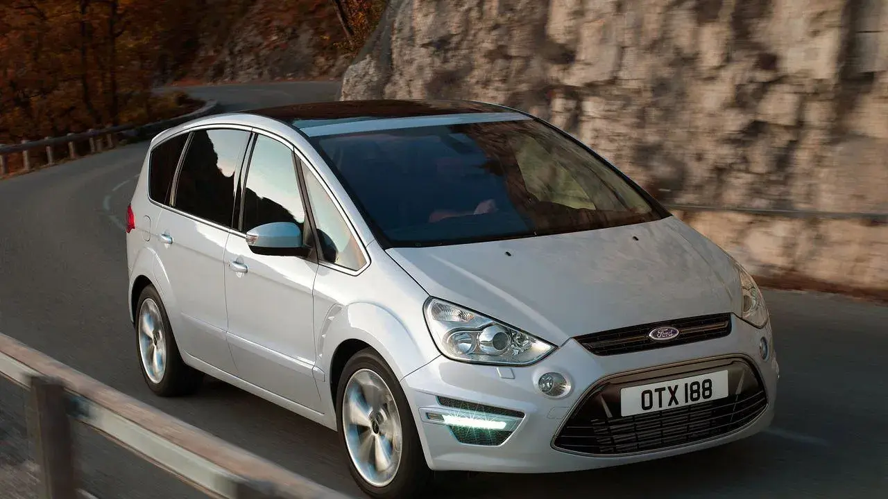 Biały Ford S-Max 2.0 TDCI pokonuje zakręt na malowniczej drodze wśród jesiennych drzew i skał.