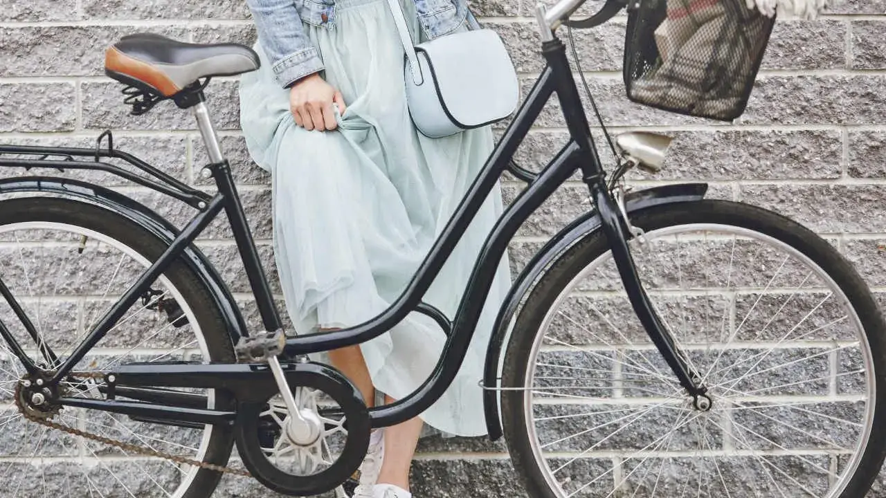 Elegancka kobieta w zwiewnej sukience i dżinsowej kurtce opiera się o rower. Widoczny rozmiar ramy roweru sugeruje, że jest to model miejski, idealny na przejażdżki po mieście.