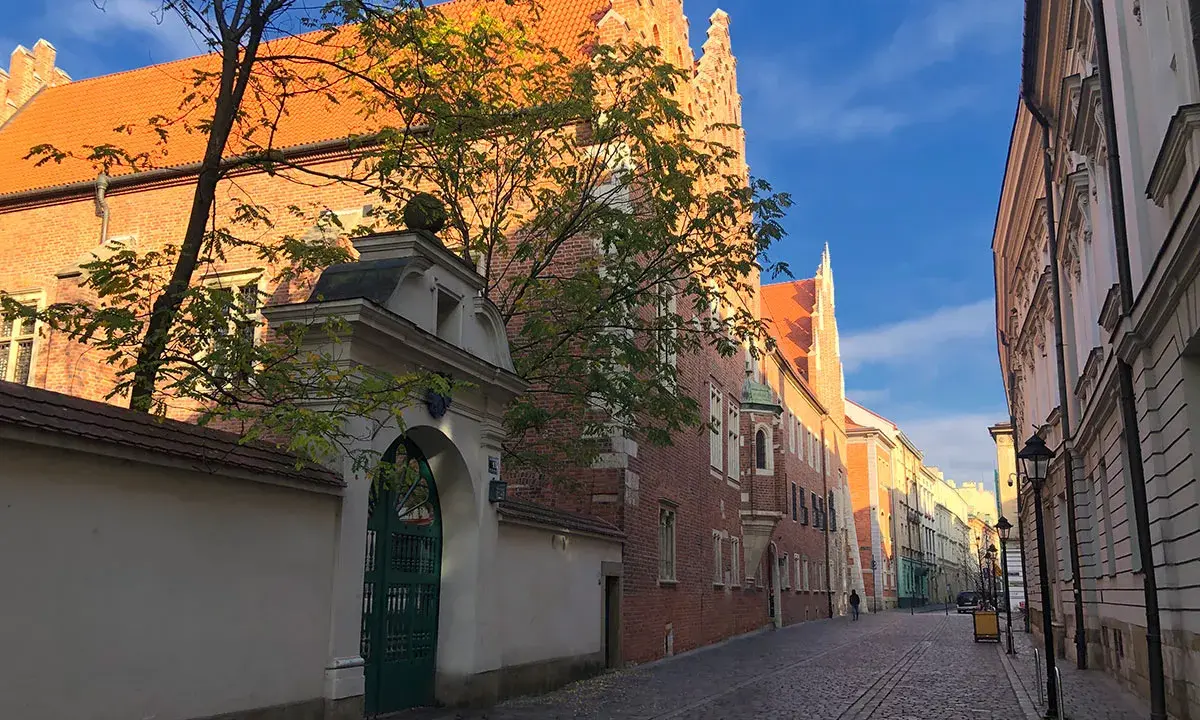 Ulica z brukowaną nawierzchnią prowadzi do zabytkowego Collegium Maius. Złote światło słońca oświetla ceglane mury i pomarańczowe dachy.