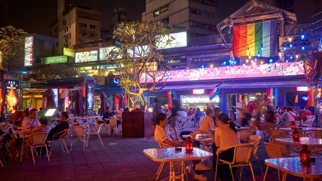 A vibrant night scene in gay Taipei, with people dining outdoors under colorful lights and a rainbow flag.