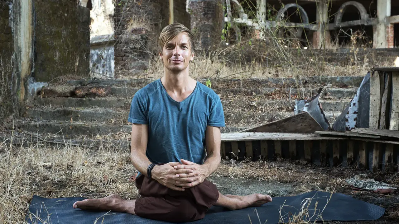 Man i blå tröja sitter i lotusställning på en yogamatta i ruiner. En stunds natthiko meditation i en övergiven miljö.