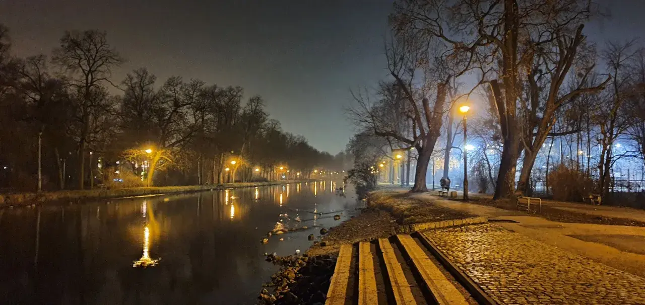Mglisty wieczór nad **Bydgoszczą**, latarnie oświetlają spokojną **rzekę** i aleję drzew.