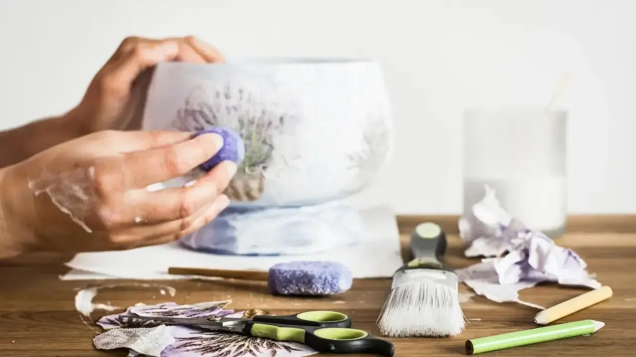 Ręce ozdabiające wazon techniką decoupage. Na stole nożyczki, pędzel, serwetki z lawendą i klej.