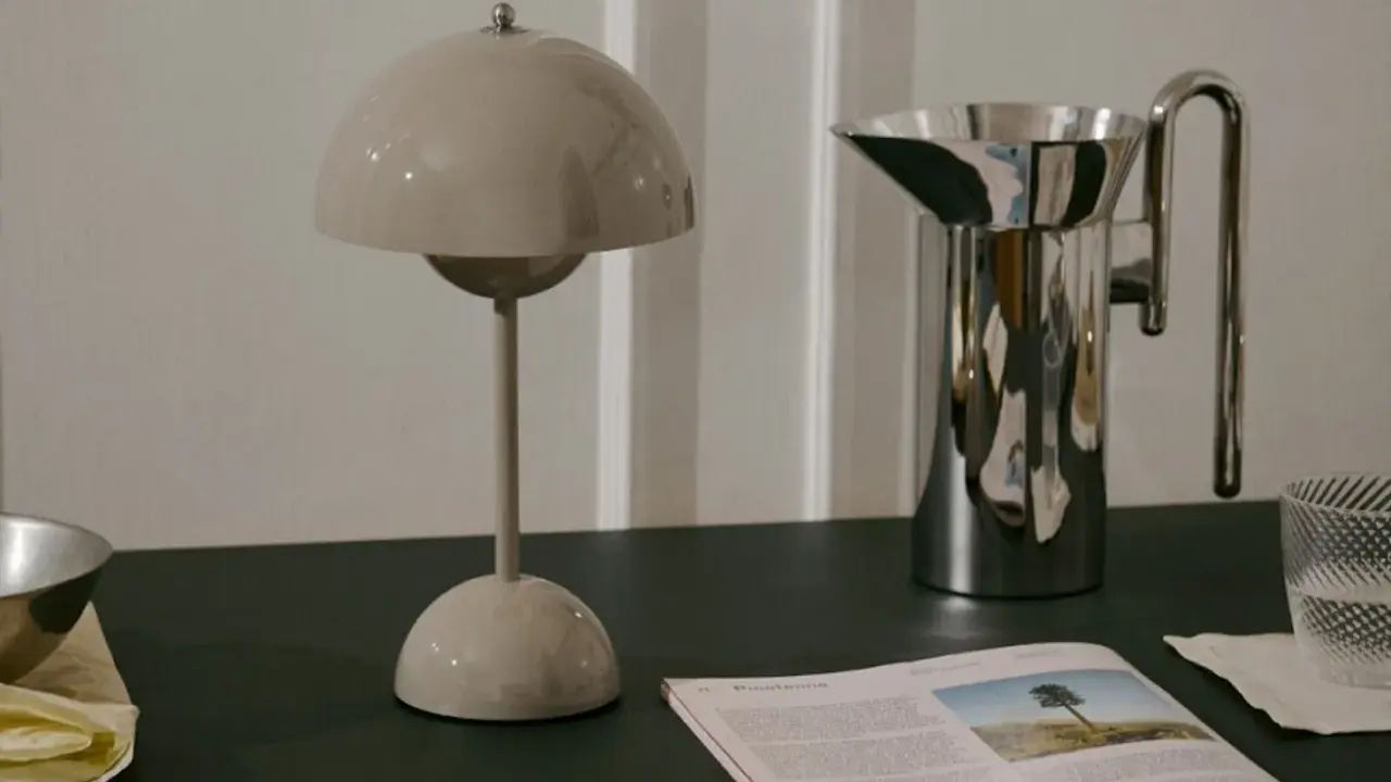 En beige lampa i studio lawahl-stil står bredvid en blank vattenkanna. En tidning med ett träd på omslaget ligger framför.