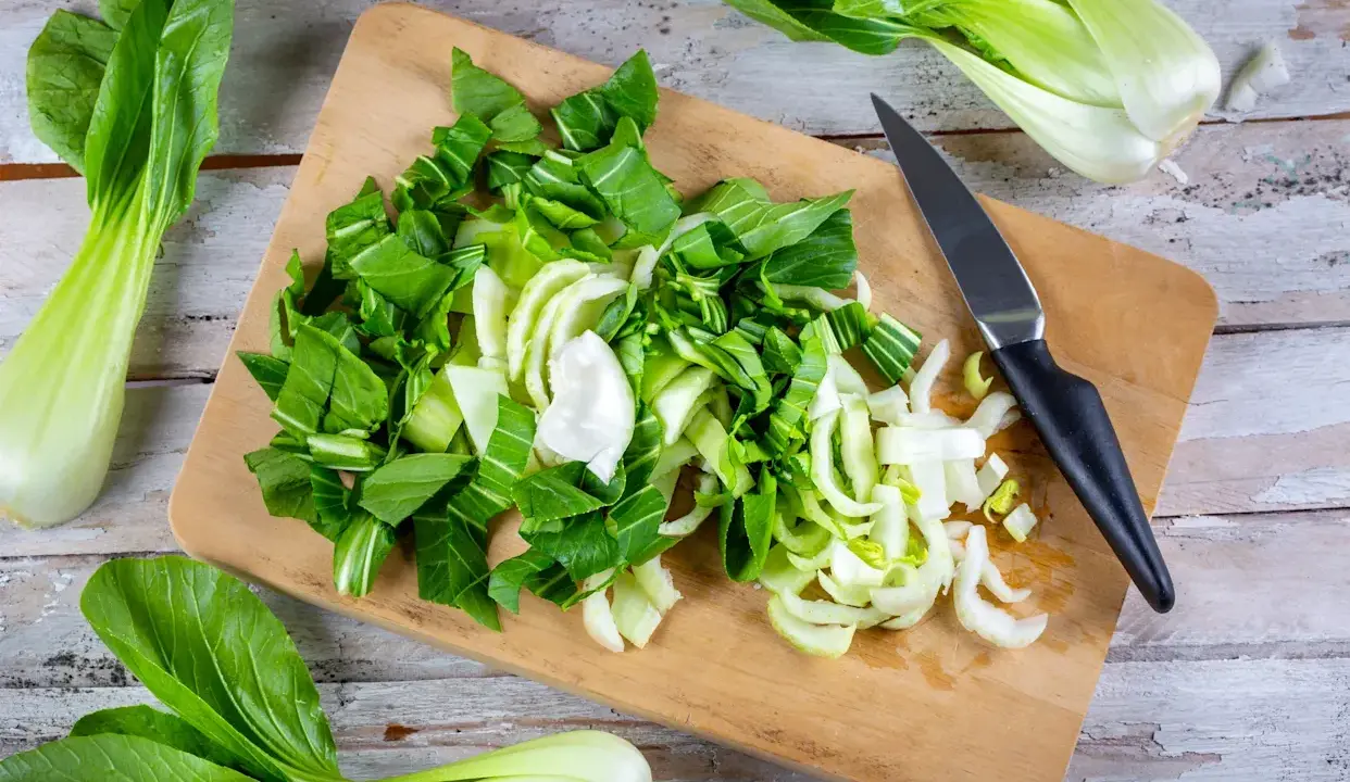 Frischer Pak Choi wird auf einem Holzbrett zerteilt, bereit zum Zubereiten. Ein Messer liegt daneben.