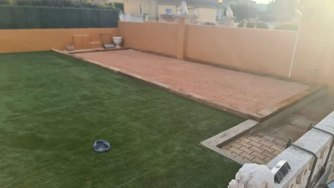 Un terrain de pétanque prêt pour l'entretien, avec une allée en gravier et une pelouse artificielle.