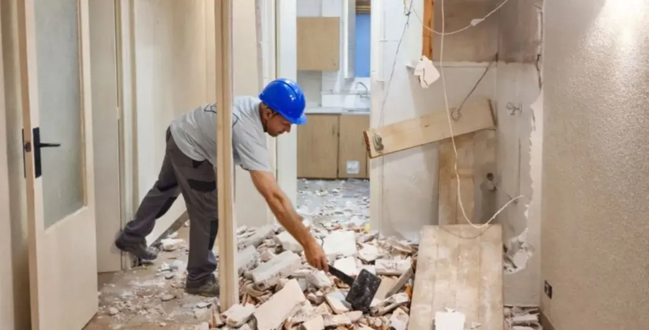 Obrero con casco azul trabajando en la demolición de un tabique, rodeado de escombros.