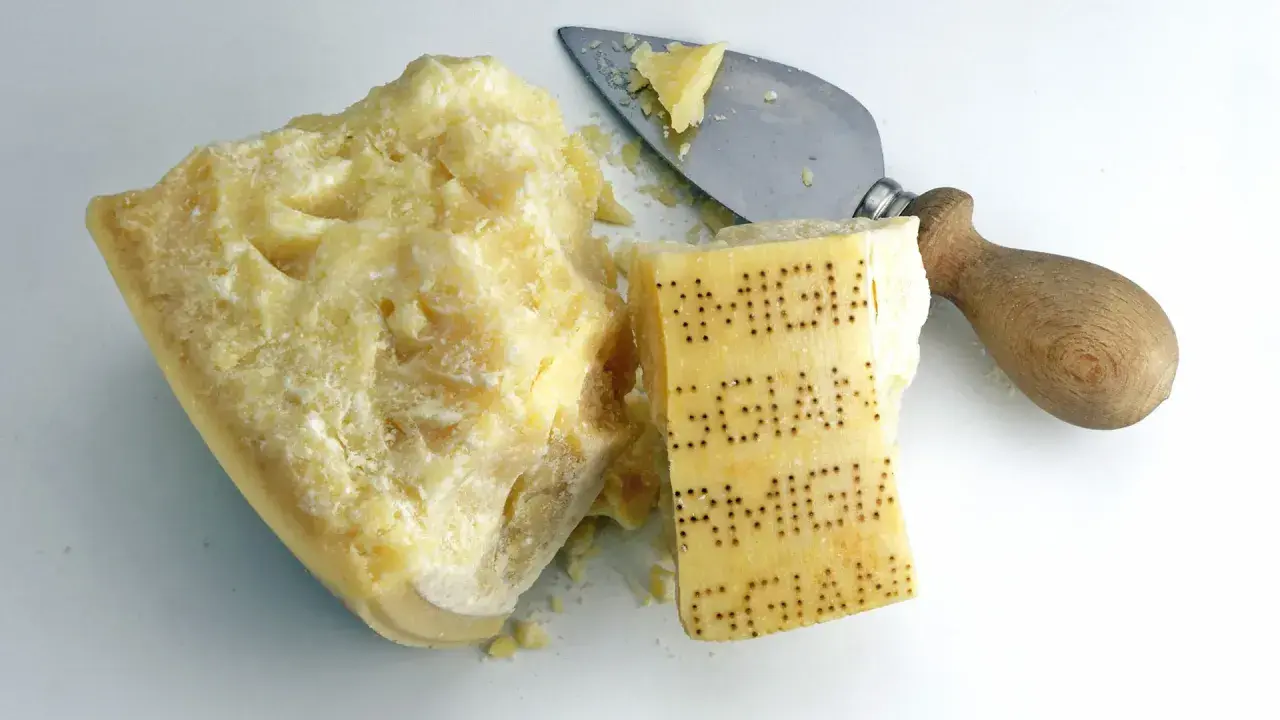 Ein Stück Parmesan mit eingeprägtem Schriftzug und ein Käsemesser. Aus was besteht Parmesan? Aus Milch, Lab und Salz.