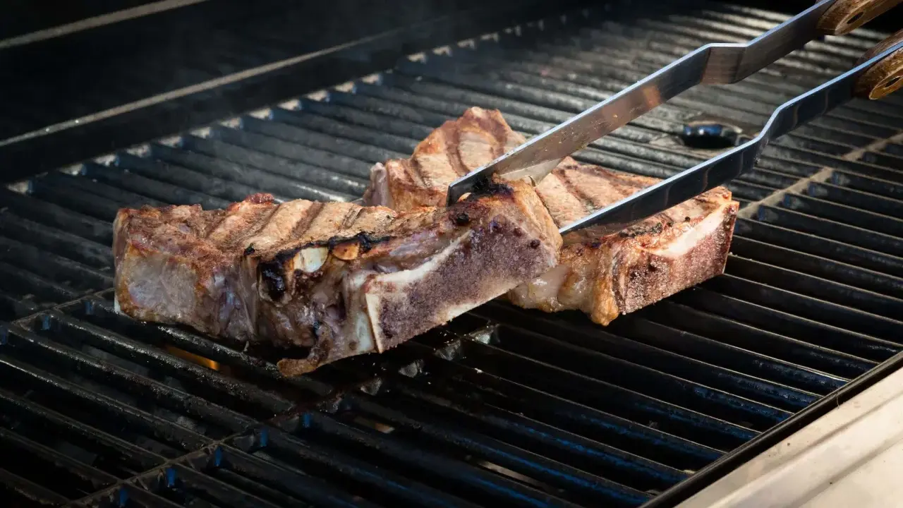 Zwei saftige Cowboy-Steaks werden mit einer Zange auf einem heißen Grill gewendet. Rauch steigt auf, während die Flammen darunter lodern.