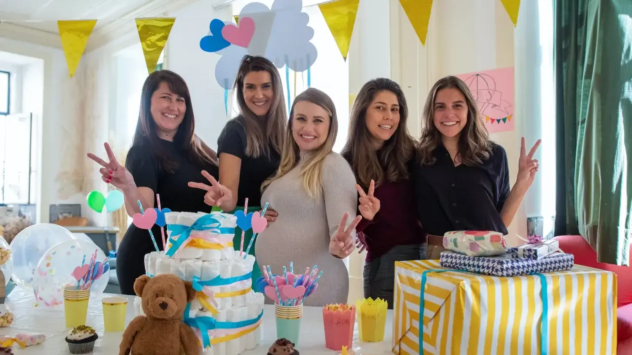 Pięć uśmiechniętych kobiet świętuje baby shower. Na stole tort z pieluch, prezenty i słodkości. To radosne wydarzenie, co to jest baby shower, pełne miłości i oczekiwania.