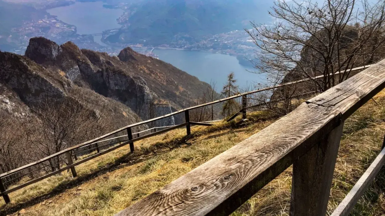 Sentiero con staccionata in legno per una passeggiata sul Pian dei Resinelli, con vista panoramica sul lago e le montagne.