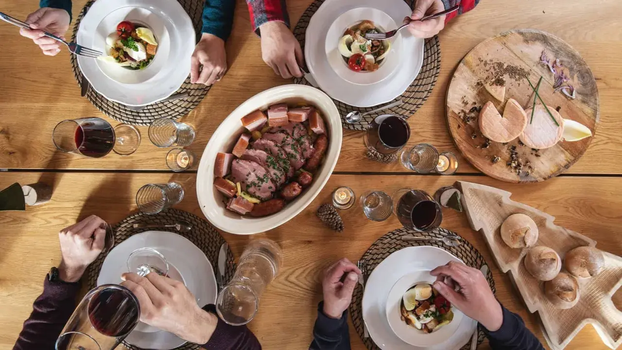 Un repas du terroir avec viande, saucisses, fromages et pain sur une table en bois. Des mains servent et dégustent.