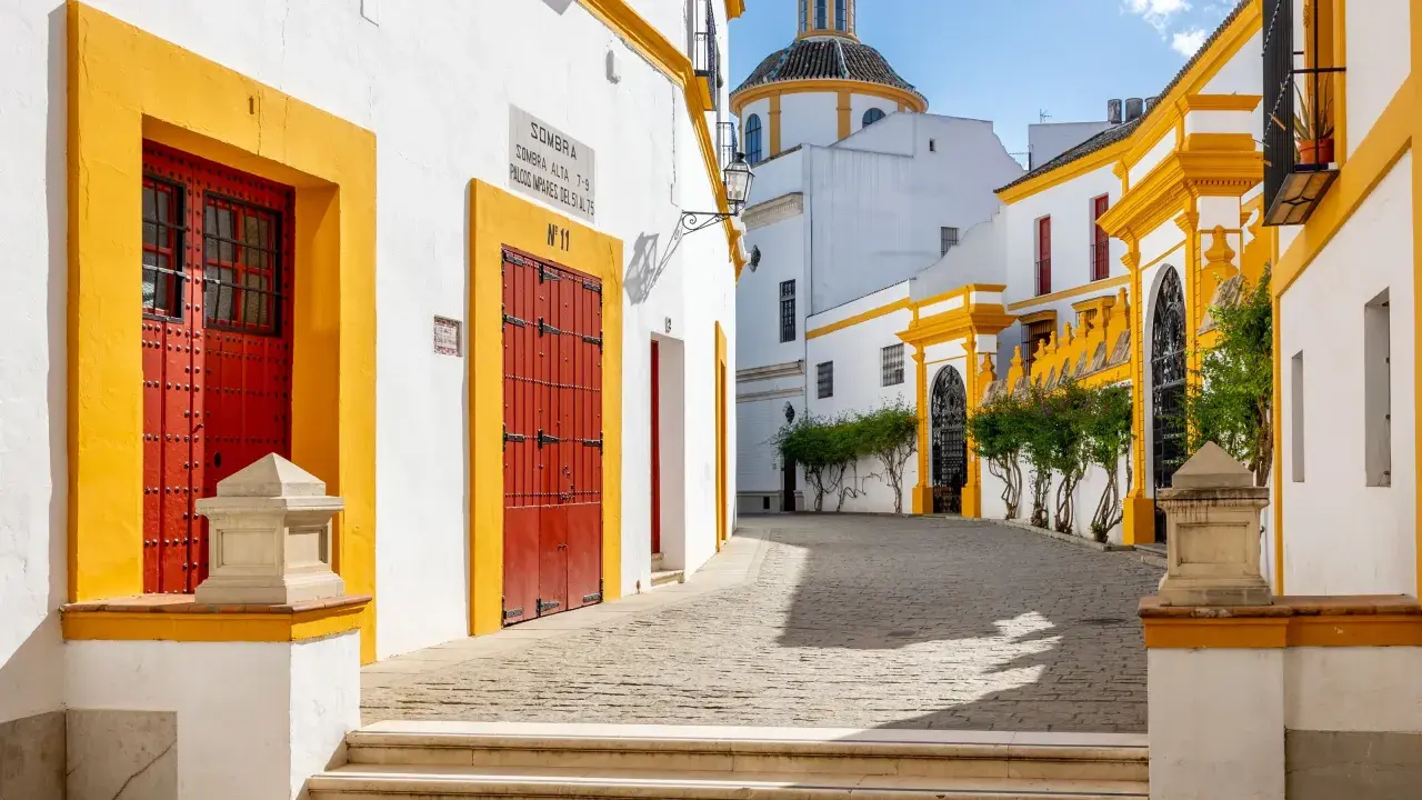 En solig gränd i Sevilla, Spanien, med vita hus, gula detaljer och röda dörrar. En kupol syns i bakgrunden.