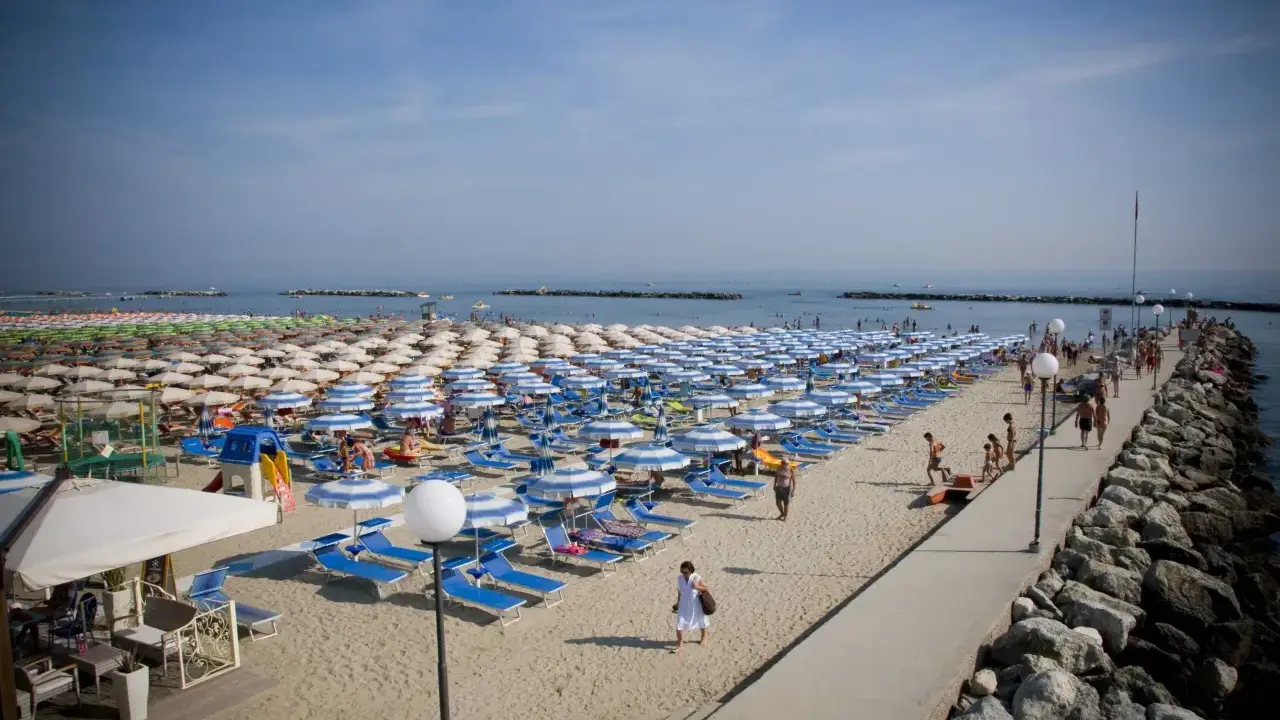 Spiaggia affollata a Rimini Gatteo Mare, con file ordinate di ombrelloni e lettini blu e bianchi, bagnanti e un molo di roccia.