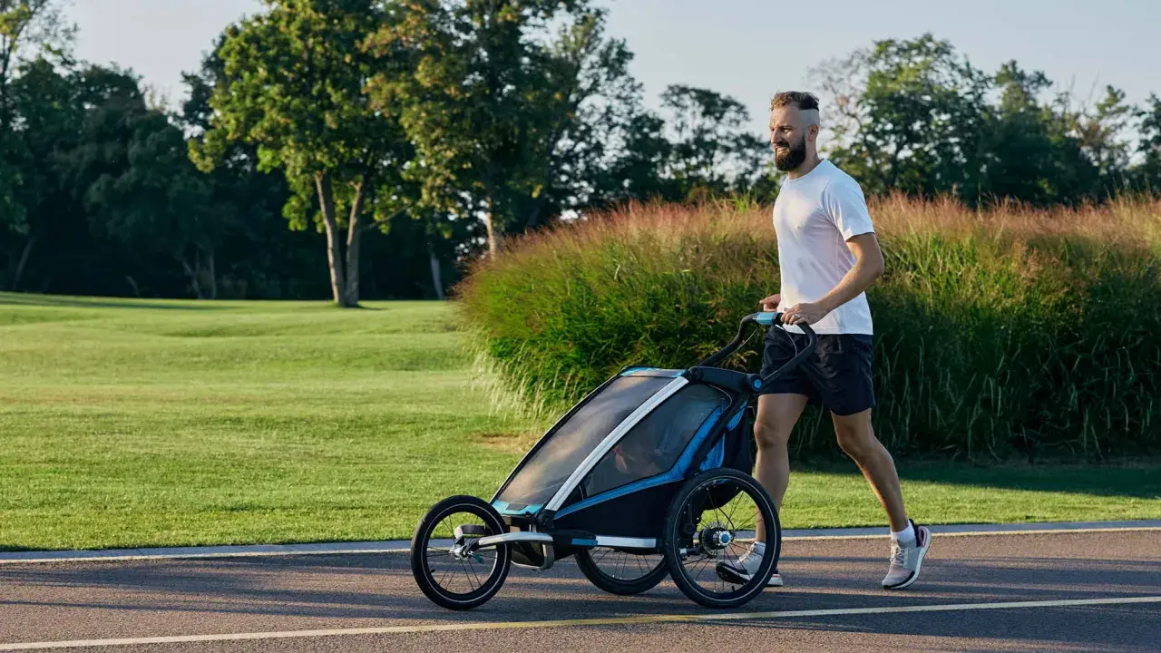 Ein Mann joggt mit Kinderwagen durch eine grüne Parklandschaft.